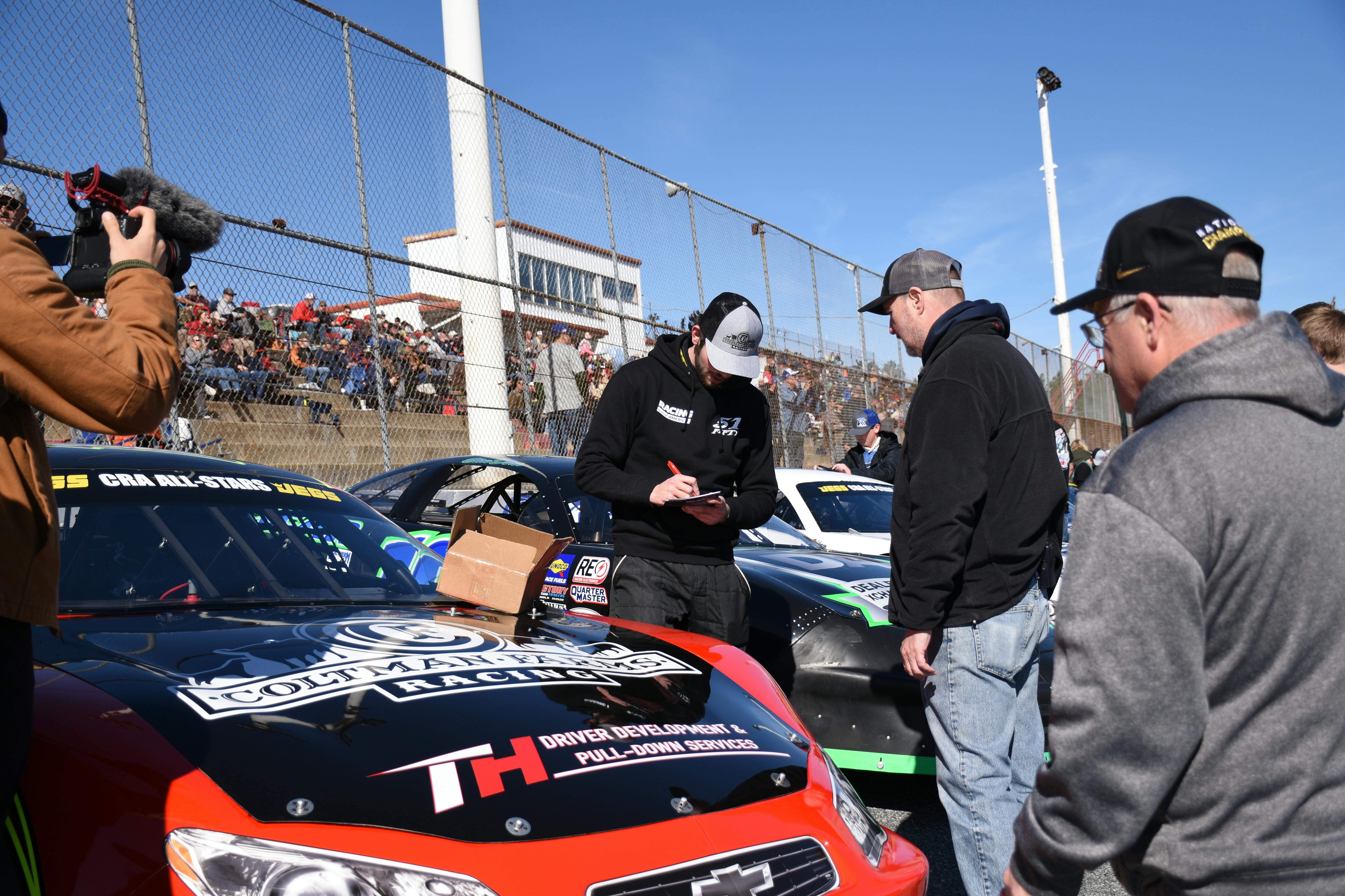 Enthusiasts gather around race cars at a motorsport event on a sunny day, engaging with drivers.